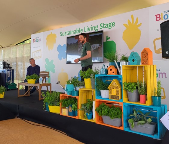 A man presents at the Bord Bia Sustainable Living Stage. On the left, another person listens attentively. The stage is sheltered underneath a large teepee tent, and various coloured wooden boxes and plants are arranged in a stacked presentation to the right of the presenter.