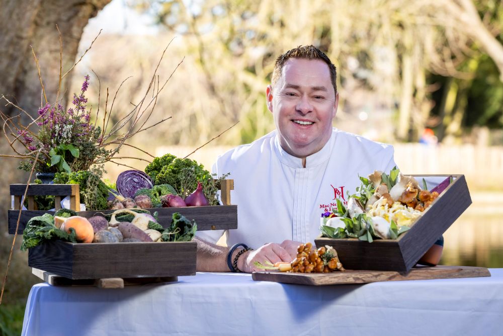 Neven Maguire poses in a n outdoor setting, smiling at the camera beside a table adorned with a variety of fresh produce.